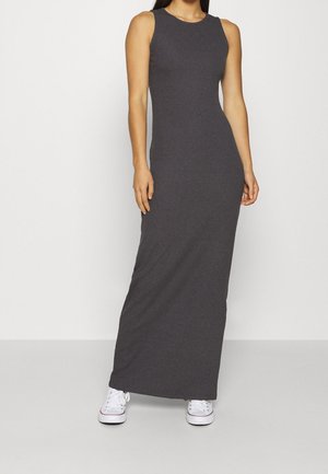 Femme portant une robe ajustée sans manches, gris foncé, longueur cheville, assortie à des baskets blanches, debout devant un fond clair uni.