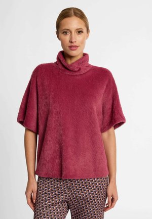 Frau trägt einen flauschigen, rosafarbenen Kurzarm-Rollkragenpullover und gemusterte braune, pinke und blaue Hosen und steht vor einem weißen Hintergrund.