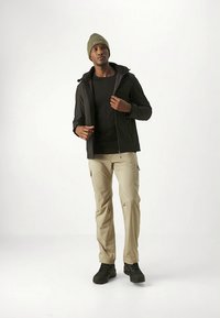 Zwarte ritsjack, zwart shirt, beige cargobroek en zwarte schoenen. Accessoires omvatten een groene gebreide muts. De stof lijkt duurzaam.