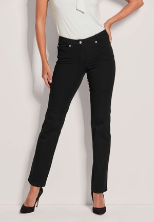 Frau in schwarzen Jeans mit geradem Bein, weißem ärmellosem Oberteil und schwarzen High Heels, stehend vor einem schlichten hellen Hintergrund.
