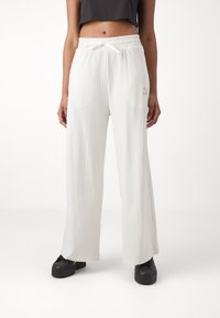 Žena v bílých žebrovaných širokých kalhotách s páskem na stažení a černých platformových botách, s krátkým černým topem viditelným nad pasem.