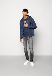 Marine gewatteerde jas met rode accenten, ritszakken, gedragen over een blauwe graphic sweatshirt; gecombineerd met versleten grijze jeans en witte sneakers.