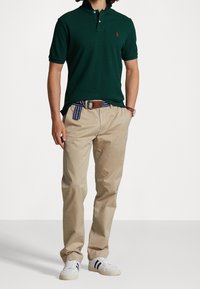 Zielona koszulka polo z dwoma guzikami, beżowe chinosy, pasek w niebiesko-żółte paski i białe sneakersy z granatowymi paskami. Model stoi przodem.