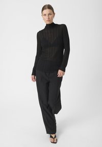 Melnā adīta turtlenecka džemperis ar ribotu faktūru, ar caurspīdīgu rakstu, kopā ar platām melnām biksēm un melniem siksnām sandalēm.