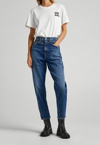Kvinna som bär en vit Pepe Jeans t-shirt, högmidjade blå jeans och svarta ankle boots, står med ena handen i fickan.