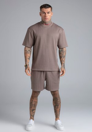 Man staat recht vooruit kijkend, draagt een taupekleurig geribd shirt met korte mouwen en bijpassende short met witte sneakers, tatoeages zichtbaar op armen en benen.