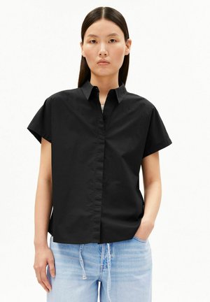 Vrouw met recht donker haar draagt een losse zwarte T-shirt met korte mouwen en lichtblauwe spijkerbroek, staand met één hand in de zak tegen een witte achtergrond.