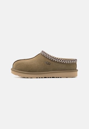 Brun ruskind slip-on tøffel med syet kant, tyk gummisål og UGG-logo på siden, vist i profil på hvid baggrund.