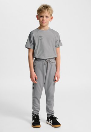 Junge steht und trägt ein graues kurzärmliges Oberteil, passende graue Jogginghosen und schwarze Turnschuhe vor einem schlichten weißen Hintergrund.