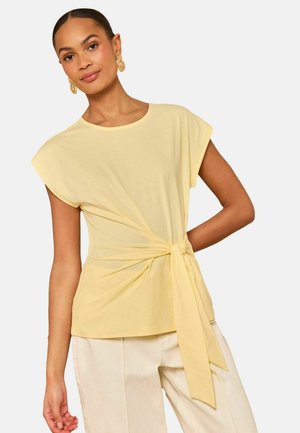 Naine, kes kannab helekollast lühikeste varrukatega ülaosa küljeotsasõlmega ja kreemivärvilisi pükse, aksessuaaridena kuldkõrvarõngad.