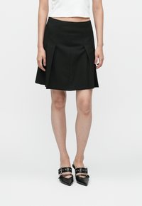 Fustă plisată neagră, cu un talie ajustată, textură netedă și tiv evazat, asortată cu pantofi negri cu vârful ascuțit și curele pentru gleznă.