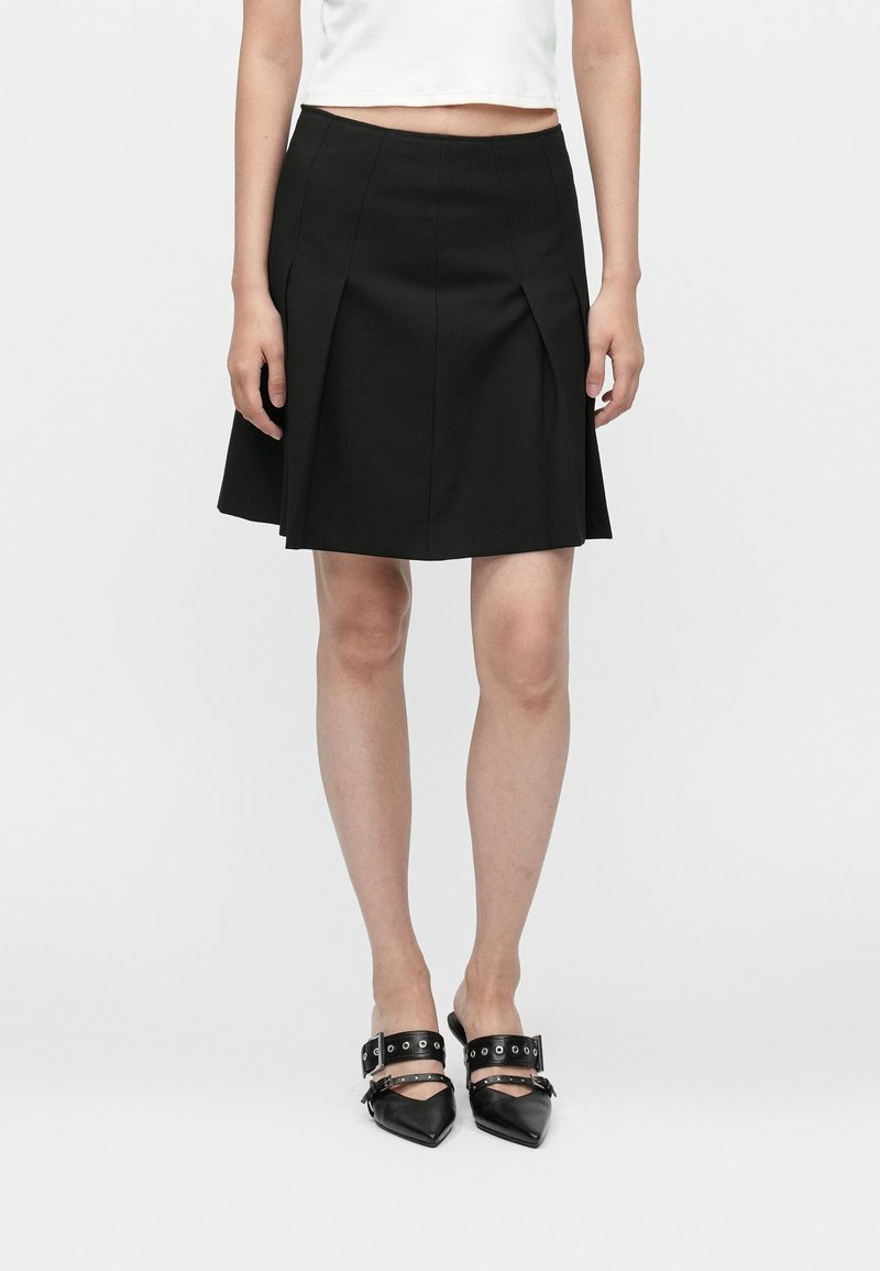 Fustă plisată neagră, cu un talie ajustată, textură netedă și tiv evazat, asortată cu pantofi negri cu vârful ascuțit și curele pentru gleznă.