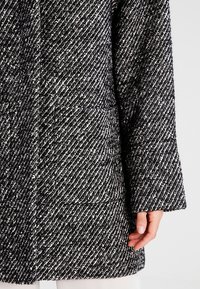 Cappotto testurizzato in bianco e nero con un motivo diagonale, dotato di maniche larghe e due tasche frontali. Il tessuto sembra spesso e caldo.