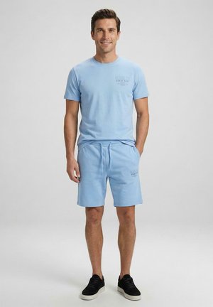 Man staand, draagt een lichtblauw T-shirt met korte mouwen en bijpassende shorts met zwarte sneakers, glimlachend tegen een effen grijze achtergrond.
