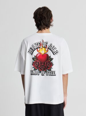 Uomo che indossa una maglietta bianca oversize con cuore ardente rosso e grafica di tre rose, testo "Cuore d'oro mani d'acciaio" sul retro.