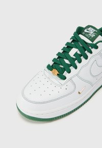 Bele usnjene superge z zelenimi vezalkami, ki imajo perforirano konico, zlate detajle na jeziku in zeleni gumijasti podplat z teksturiranimi poudarki.