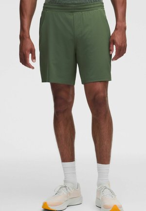 Hombre de pie, vistiendo pantalones cortos deportivos verde oliva, calcetines blancos hasta el tobillo y zapatillas blancas con detalles naranjas.