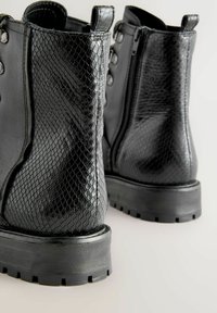 Fekete bokacsizma, az egyik oldalán texturált kígyóbőr mintázattal, a másikon sima bőrrel, vastag talppal és oldalsó cipzárral.
