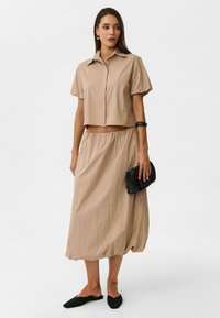Beige cropped shirt met korte pofmouwen en knoopsluiting, gecombineerd met een bijpassende mid-length rok met elastische tailleband. Zwarte clutch en schoenen.