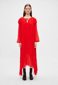 Vestido de gasa roja con mangas largas abullonadas, acentos con volantes y un dobladillo asimétrico. Presenta un escote en forma de ojal y una textura suave.