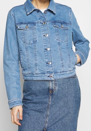 Frau trägt eine hellblaue Jeansjacke mit Metallknöpfen und zwei Brusttaschen, kombiniert mit einem passenden Jeansrock.