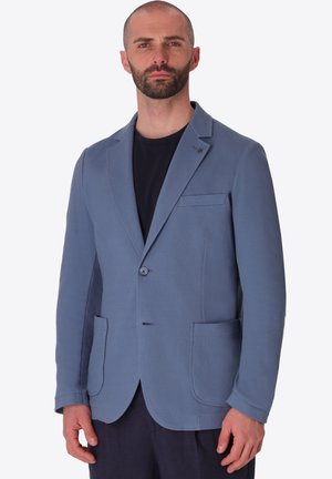 Uomo con barba corta che indossa un blazer azzurro chiaro strutturato sopra una camicia nera, in piedi con espressione neutra su uno sfondo semplice.