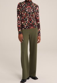 Halenka s dlouhým rukávem a vysokým výstřihem s barevným abstraktním vzorem v červené, zelené a béžové barvě, spárovaná s olivově zelenými širokými kalhotami a černými botami.