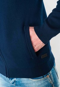 Marineblauwe zip-up sweater met een zachte textuur, voorzien van een voorzak en geribbelde manchetten, gecombineerd met lichtblauwe denim jeans.