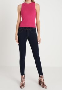 Top sin mangas rosa ajustado, jeans oscuros ajustados y botas de tobillo a cuadros con tacón grueso. Tela suave y patrones contrastantes.