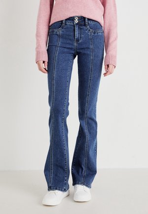 Blue denim flared jeans with contrasting stitching, high waist, and two-button closure; paired with a pink knit sweater and white sneakers.