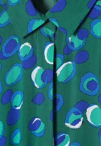 Camicia verde con motivi circolari astratti blu, turchese e bianchi, caratterizzata da un colletto classico e una cucitura frontale visibile.