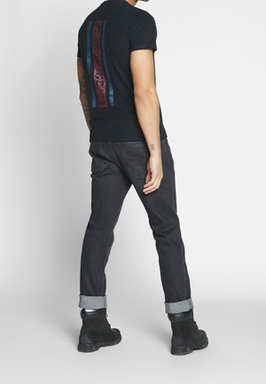 Camiseta negra con un gráfico vertical en rojo y azul, combinada con jeans de mezclilla oscura y botas negras de cordones. Estilo ajustado, con puños enrollados.