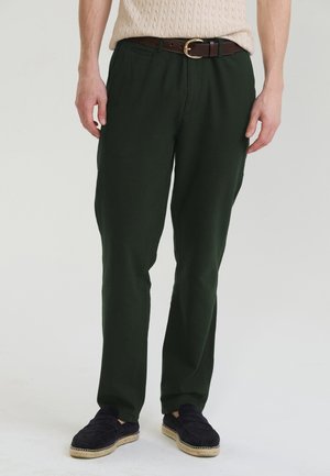 Mann trägt dunkelgrüne gerade geschnittene Hose, braunen Gürtel, beiges gestricktes Oberteil und schwarze Slip-On-Schuhe mit hellen Sohlen.