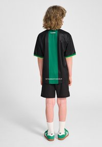 Zwarte sporttrui met verticale groene streep, korte mouwen en de tekst "INTERNATIONALS" op de achterkant, gecombineerd met zwarte shorts en groene schoenen.
