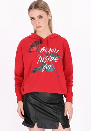 Młoda kobieta ubrana w czerwoną bluzę z kapturem z napisem „Beauty Inside Out” i grafiką róży, połączoną z czarną skórzaną mini spódniczką.