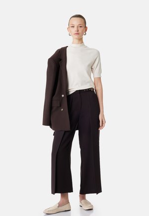 Kobieta ubrana w biały top z krótkim rękawem, czarne szerokie spodnie, trzymająca czarną marynarkę na jednym ramieniu, z beżowymi mokasynami i kolczykami kołami.