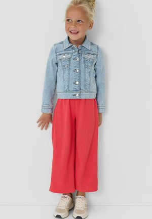 Lachend meisje met blond haar in een knot, draagt een lichtblauwe spijkerjack, rode wijd uitlopende broek en beige sneakers, staand tegen een witte muur.