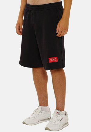 Schwarze Baumwollshorts mit lockerer Passform, elastischem Bund und einem roten Label-Patch am linken Bein. Tragen mit weißen Sneakers.