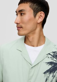 Lichtgroen kortemouwenshirt in een ontspannen pasvorm, met een donkere palmbomenontwerp aan de linkerkant. Witte t-shirt eronder gelaagd.