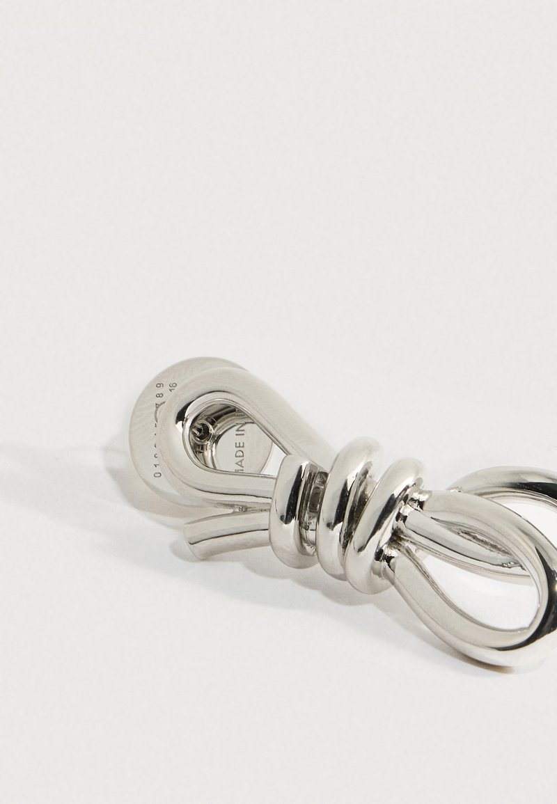 Accesorio brillante de metal plateado con forma de nudo, colocado sobre una superficie blanca, con texto grabado parcialmente visible en un extremo.