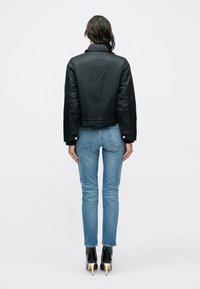 Γυναίκα με πλάτη, φοράει μαύρο cropped σακάκι, ανοιχτό μπλε στενά τζιν και μαύρα μπεζετά ψηλοτάκουνα μποτάκια, μπροστά σε λευκό φόντο.