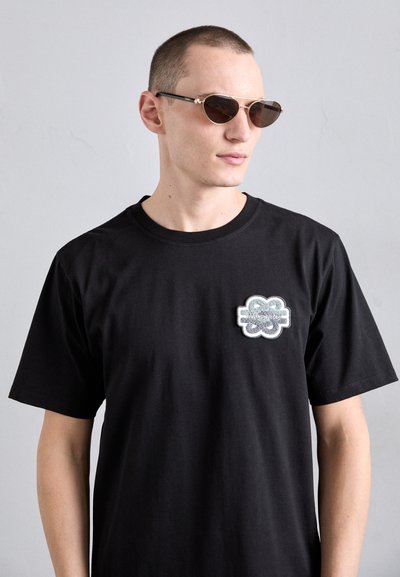 T-shirt en coton noir à manches courtes, arborant un logo en patch texturé gris sur la poitrine gauche. Le modèle porte des lunettes de soleil sombres avec des accents dorés.