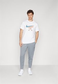 Herreoutfit med en hvid bomuldstrøje med et farverigt grafisk motiv, parret med lysegrå joggers og hvide sneakers.