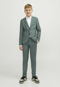 Completo color teal chiaro con giacca aderente, pantaloni coordinati e camicia bianca. Abbinato a sneakers nere con dettagli bianchi.