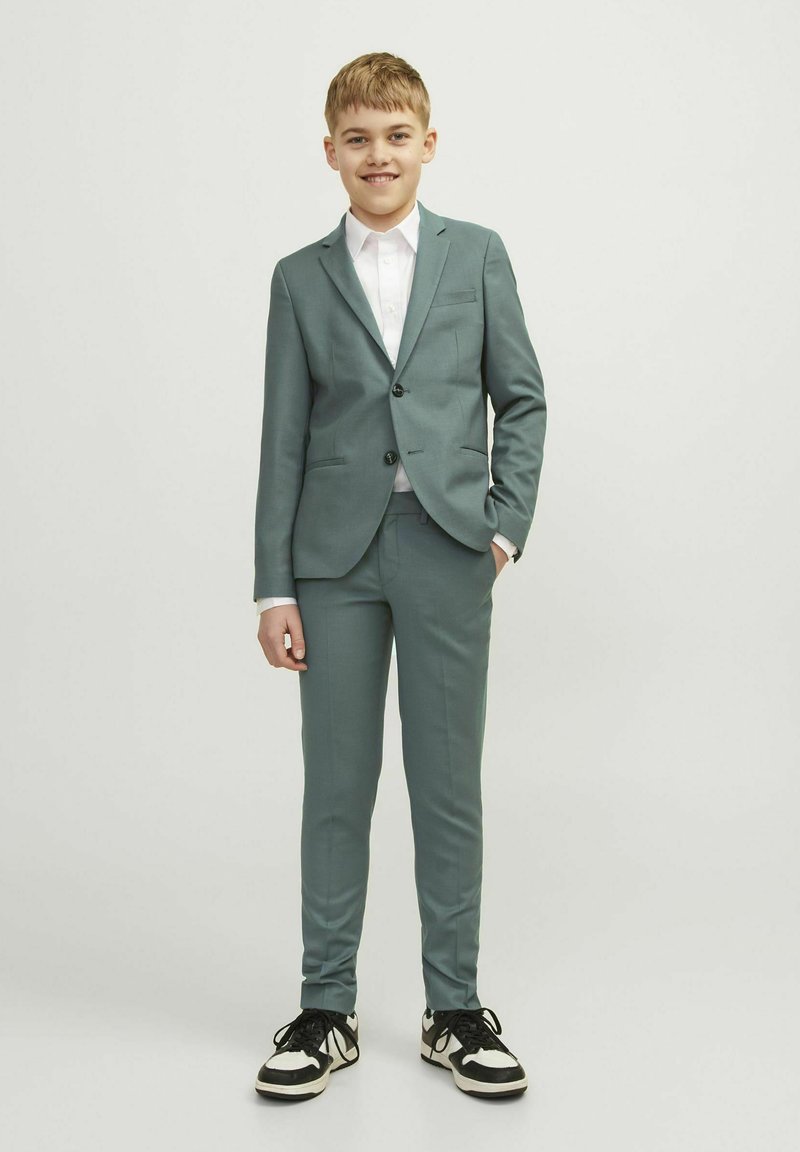 Completo color teal chiaro con giacca aderente, pantaloni coordinati e camicia bianca. Abbinato a sneakers nere con dettagli bianchi.