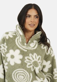 Pullover in pile verde con zip, caratterizzato da motivi floreali bianchi e spirali. Tessuto morbido e testurizzato, colletto alto e vestibilità rilassata per il massimo comfort.
