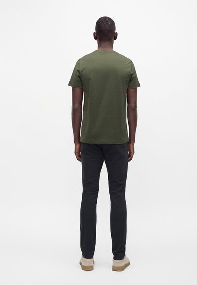 Homme portant un t-shirt vert olive à manches courtes, un pantalon noir et des chaussures beiges, debout de dos sur un fond blanc uni.