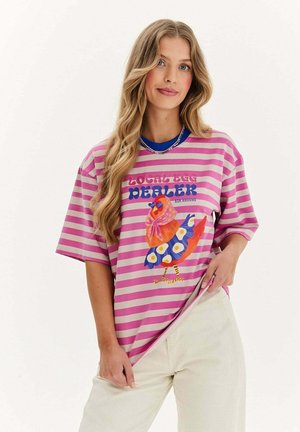 Kobieta o długich blond włosach, ubrana w różowo-fioletowy pasiasty t-shirt z kolorową grafiką kurczaka i kremowe spodnie.