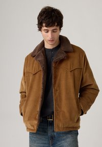 Chaqueta de pana marrón con un collar de forro polar marrón, con bolsillos frontales y un corte relajado, combinada con una camiseta oscura y jeans.