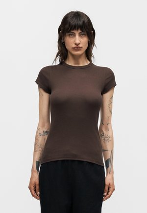 Femme aux cheveux foncés et tatouages sur les deux bras porte un t-shirt marron ajusté à manches courtes et un pantalon noir sur un fond clair uni.